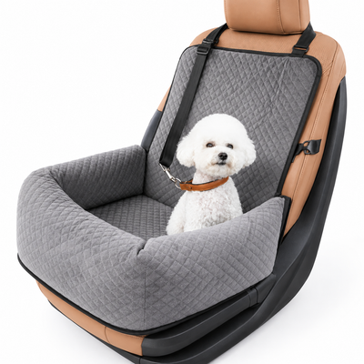 A small white dog sits inside a padded car seat cover designed for pets, shown on a clean white background. The cover is gray quilted fabric with black straps and trim, installed over a tan and black car seat.